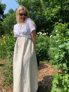 Woman stands in flower garden wearing a beige linen maxi dress and white short sleeve wrap top tied in the back.