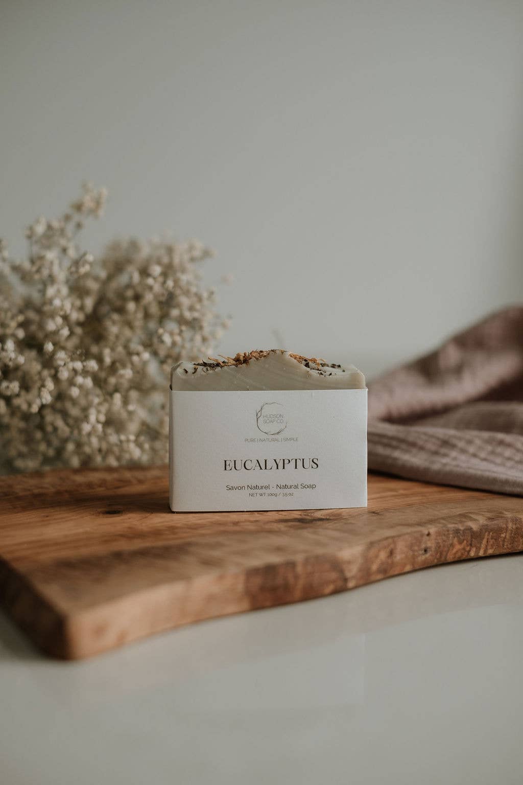 Bar of soap labeled 'Eucalyptus' on a wooden surface with dried flowers and a towel in the background.