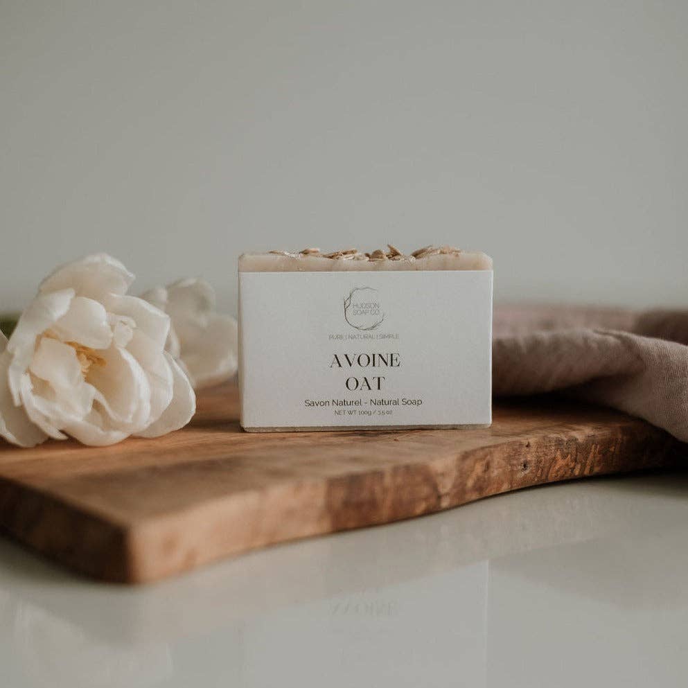 Bar of soap labeled 'Avoine Oat' on a wooden board with a white flower and brown towel in the background.