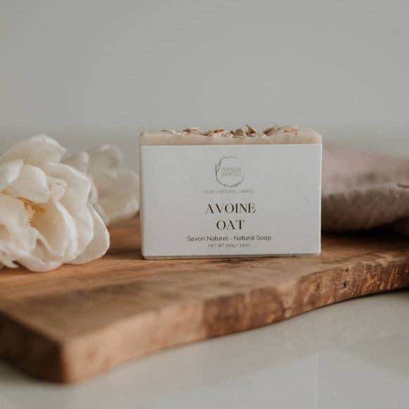 Bar of soap labeled 'Avoine Oat' on a wooden surface with a white flower in the background.