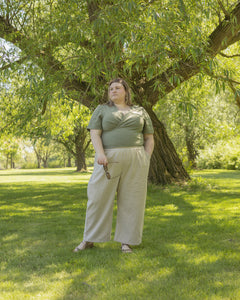 Plus size model wears a green, short sleeve wrap top and beige wide leg pants. She is standing in front of a large tree with lots of greenery around.