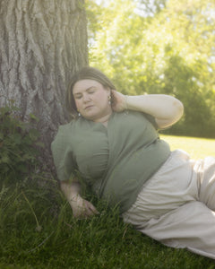 Model lays in grass wearing a green linen wrap top and loose beige pants.