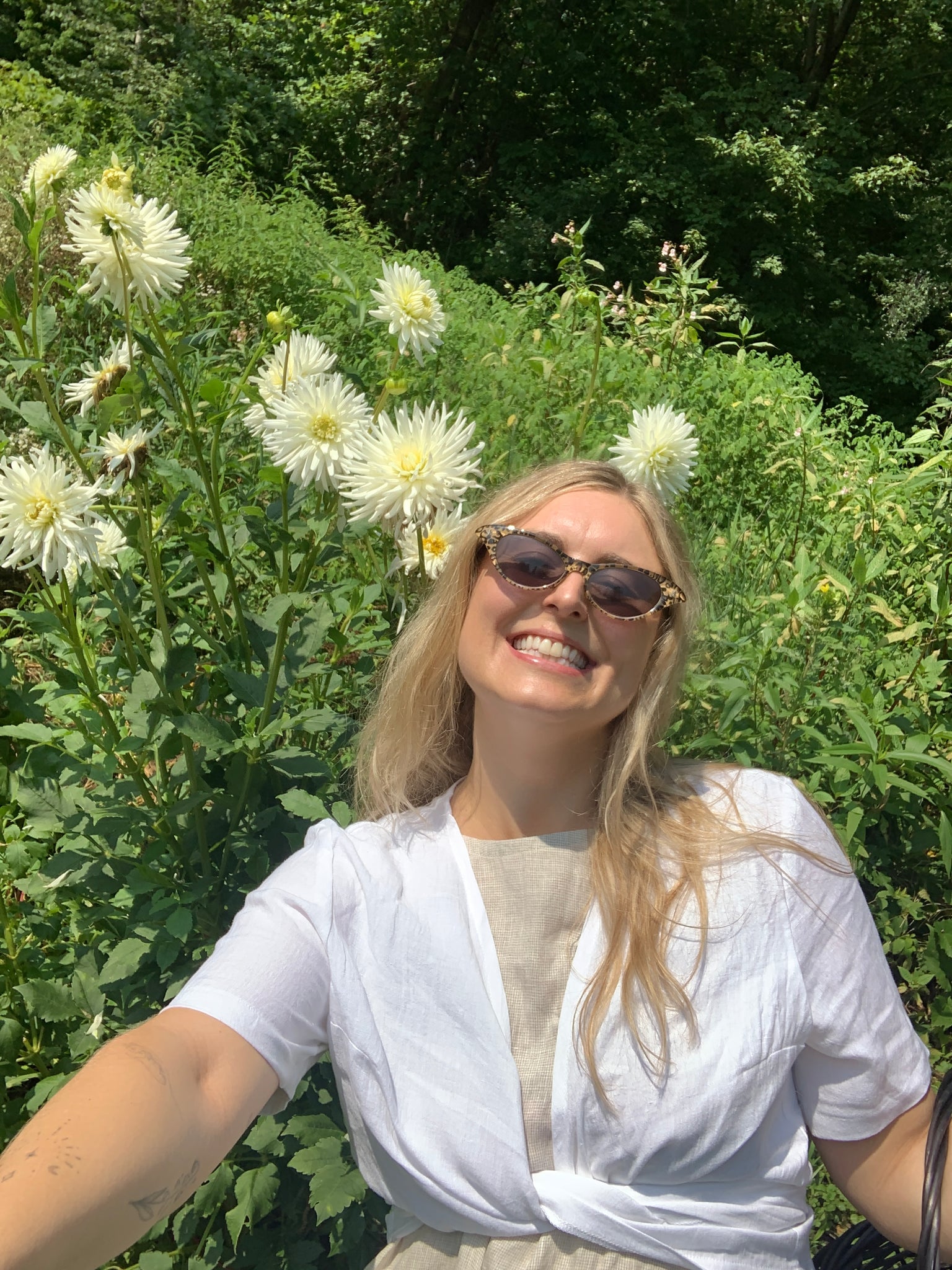 Woman wears a white linen wrap top with short sleeves and smiles next to a white dahlia flower.