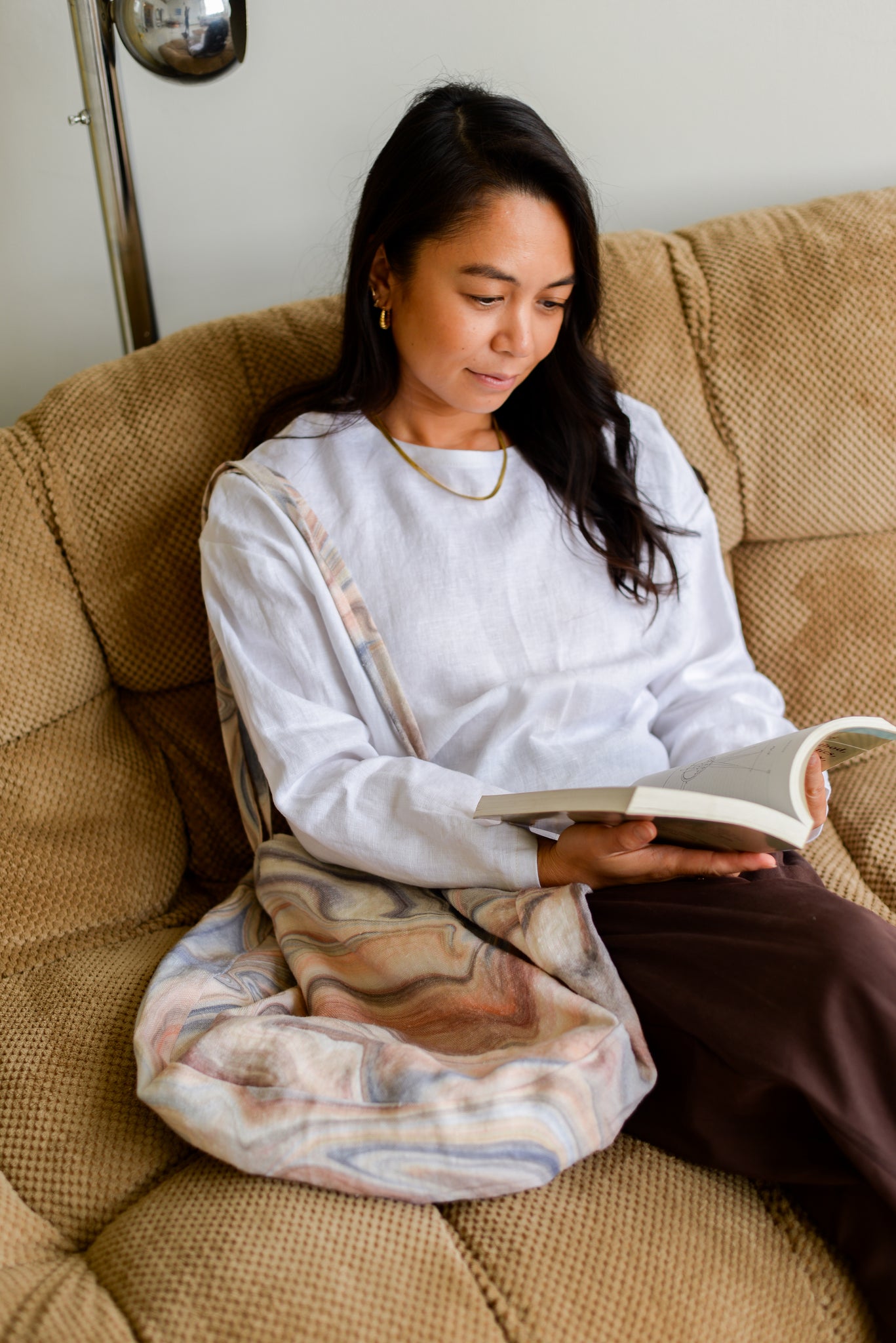 woman sitting on a couch reading wearing a white linen top and brown pants with a large printed tote